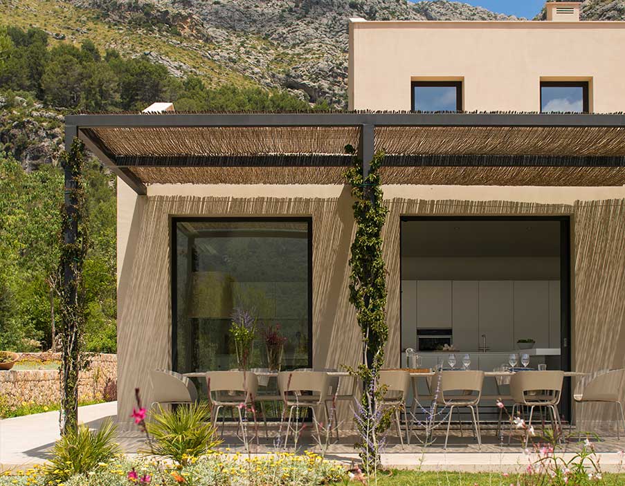 Schilfrohr-Pergola und Putzfassade mit sichtbarer weißer Küche, dahinter ein Berghang