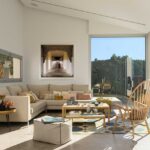 Salón luminoso con sofá esquinero beige, mesas de centro de madera, alfombra geométrica y grandes ventanales