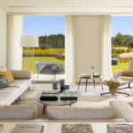 Salón con sofás de tela beige, gran cuadro azul y verde, cortinas blancas y jardín al fondo