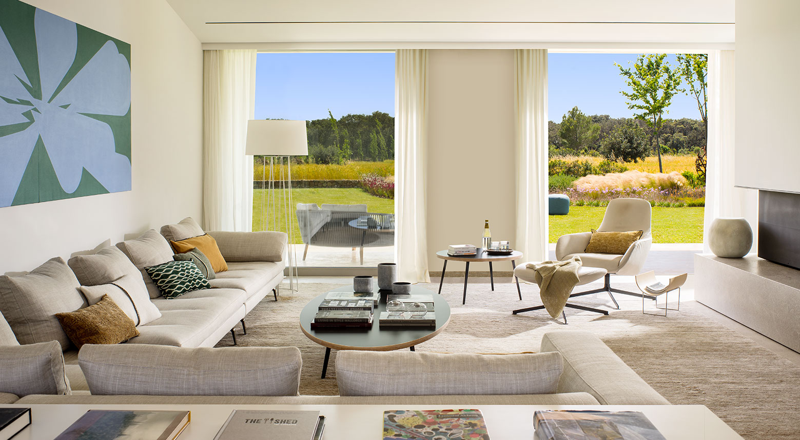 Salón con sofás de tela beige, gran cuadro azul y verde, cortinas blancas y jardín al fondo