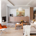 Salón luminoso con sofá beige, sillón naranja, aparador de madera clara, chimenea y suelo pulido claro