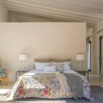 Geräumiges Schlafzimmer mit geblümter Tagesdecke, weißen Lampen, Holzsessel und weißer Balkendecke