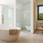 Baño con bañera exenta blanca, ducha de mármol tras mampara de cristal y suelo de piedra beige.