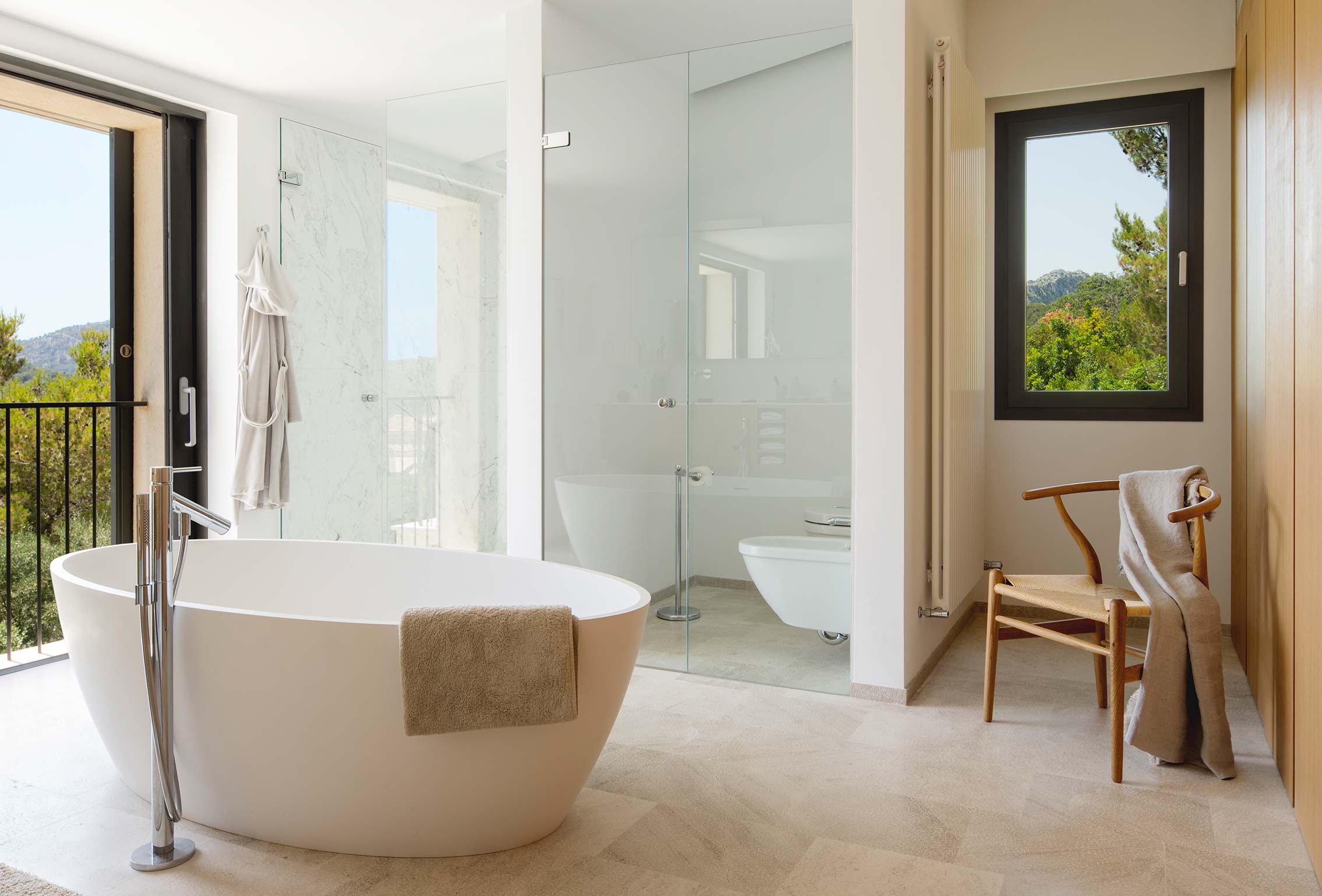 Baño con bañera exenta blanca, ducha de mármol tras mampara de cristal y suelo de piedra beige.