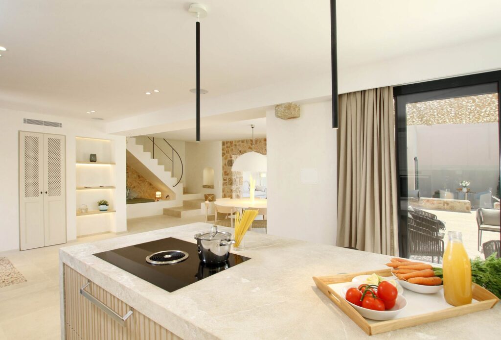 Isla de cocina de mármol beige con placa negra, escalera y comedor al fondo en planta abierta