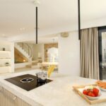 Isla de cocina de mármol beige con placa negra, escalera y comedor al fondo en planta abierta