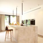 Cocina blanca con isla de mármol beige veteado, taburete de madera y cortinas junto al jardín