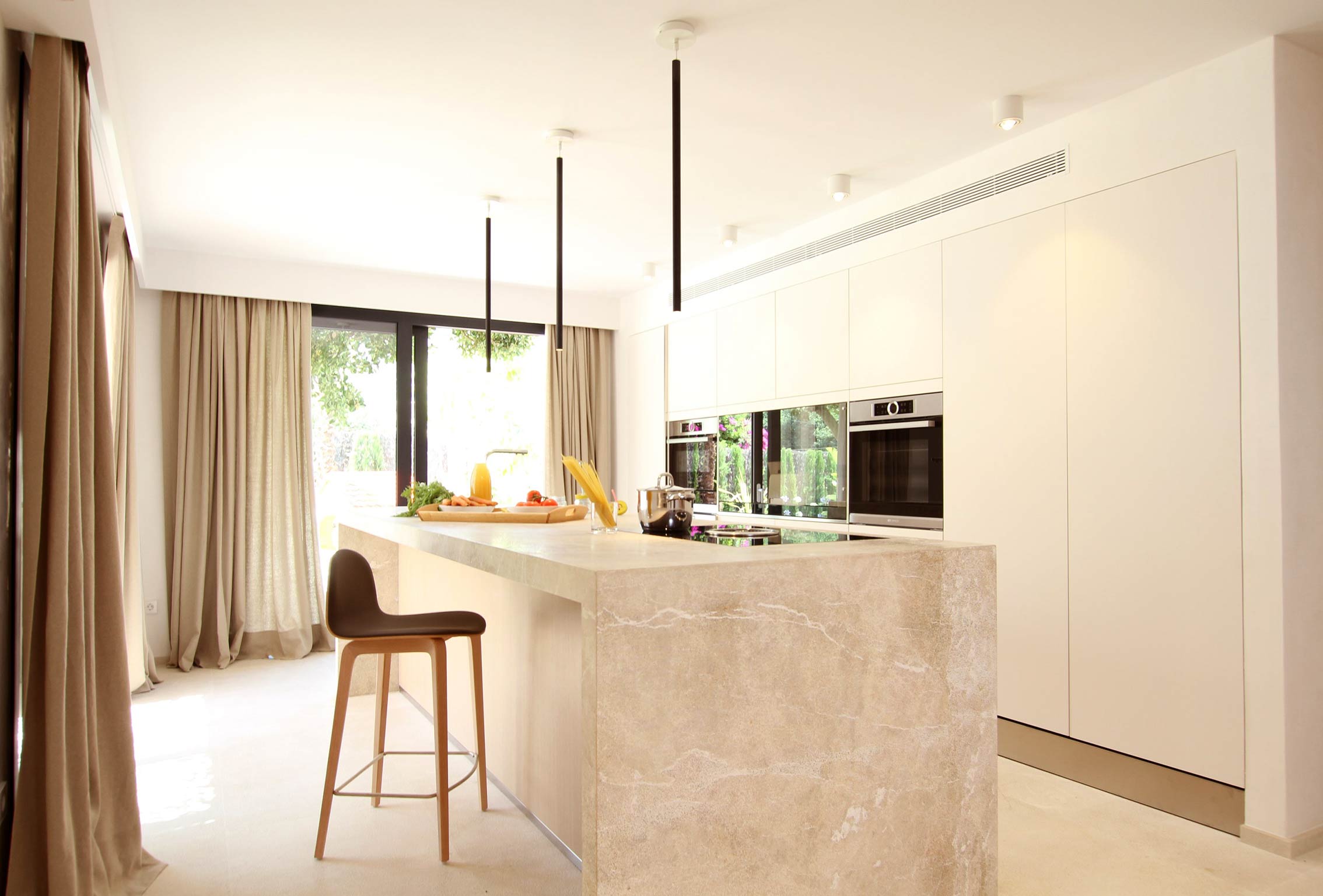 Cocina blanca con isla de mármol beige veteado, taburete de madera y cortinas junto al jardín