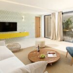 Salón con sofá blanco, sillón azul, mesa baja redonda de madera y mueble amarillo bajo la televisión