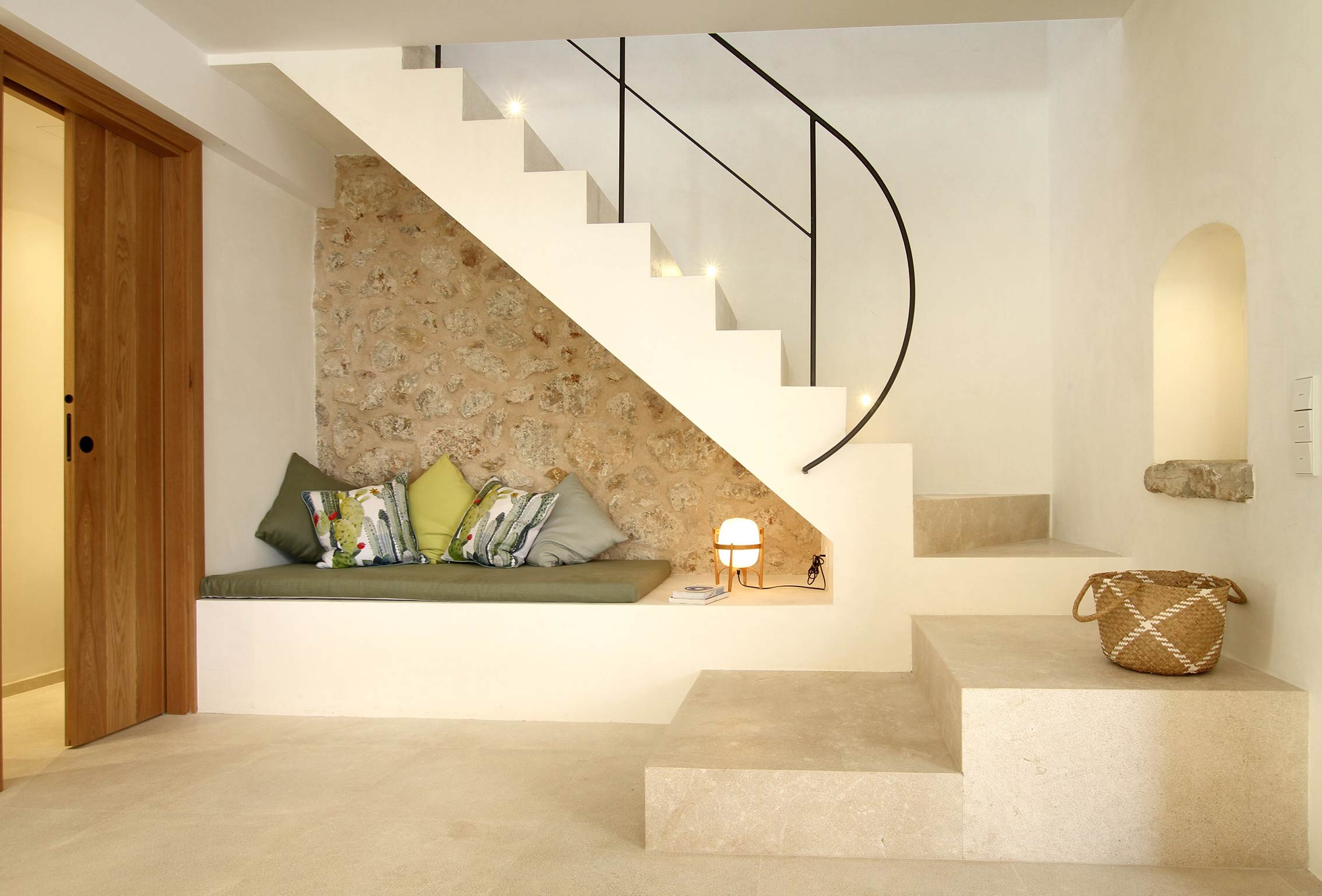 Escalera blanca con banco y cojines verdes bajo pared de piedra vista, con peldaños de piedra clara