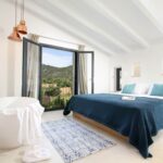 Dormitorio con vigas blancas, bañera exenta, cama con colcha azul y vistas a la montaña por la balconera