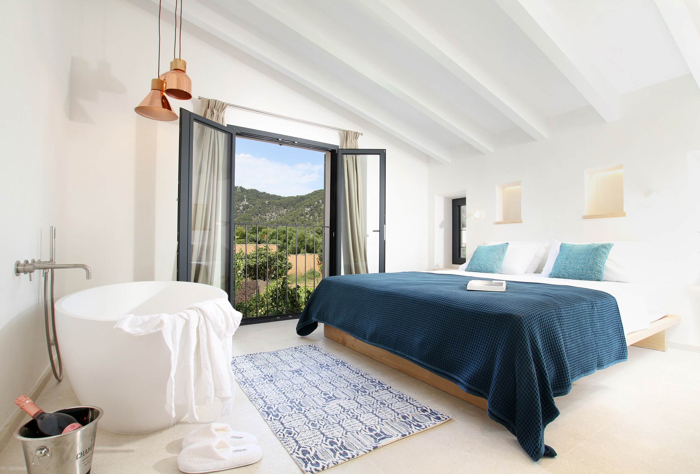 Dormitorio con vigas blancas, bañera exenta, cama con colcha azul y vistas a la montaña por la balconera