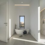 Baño con bañera exenta blanca, ducha revestida en mármol veteado y luz natural desde una ventana estrecha