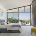 Dormitorio con cabecero azul grisáceo, sillón amarillo y gran ventanal con vistas al mar