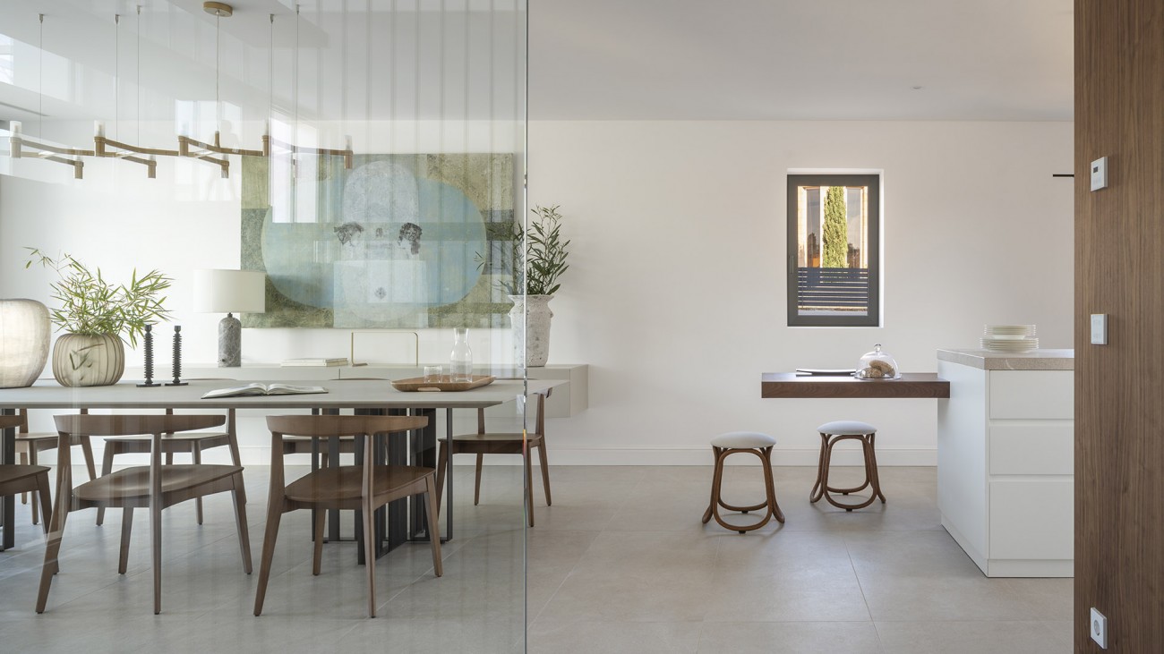 Comedor con sillas de madera visto a través de un cristal, con la cocina y taburetes al fondo