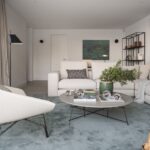 Salón con sofá crema en L, mesa de centro redonda, alfombra azulada y butaca blanca