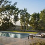 Piscina y tumbonas con luz dorada del atardecer, rodeadas de pinos, césped y muro de piedra seca