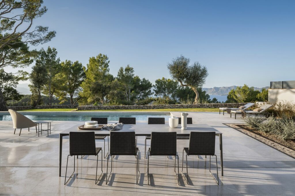 Mesa de comedor exterior con sillas oscuras junto a la piscina y vistas al mar entre pinos