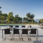 Mesa de comedor exterior con sillas oscuras junto a la piscina y vistas al mar entre pinos