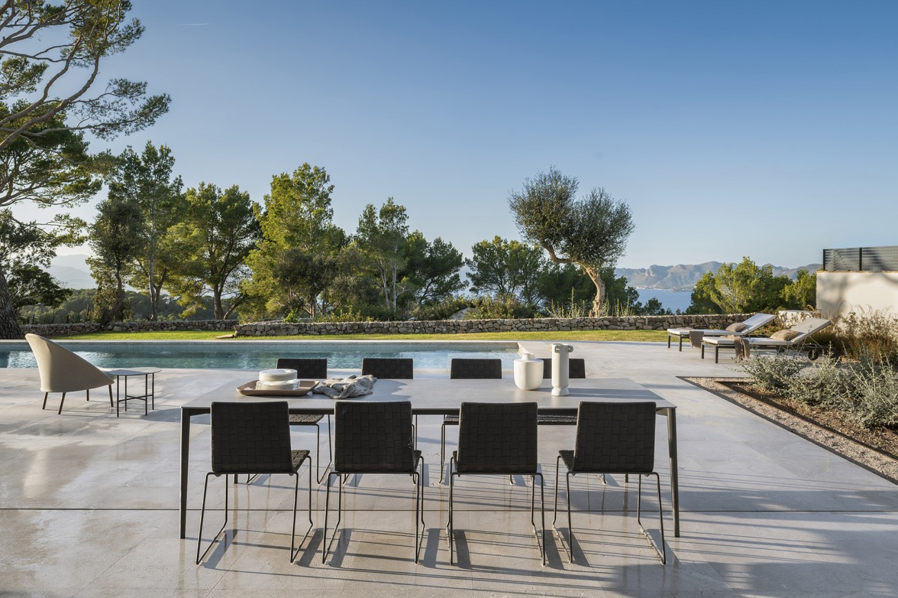 Mesa de comedor exterior con sillas oscuras junto a la piscina y vistas al mar entre pinos