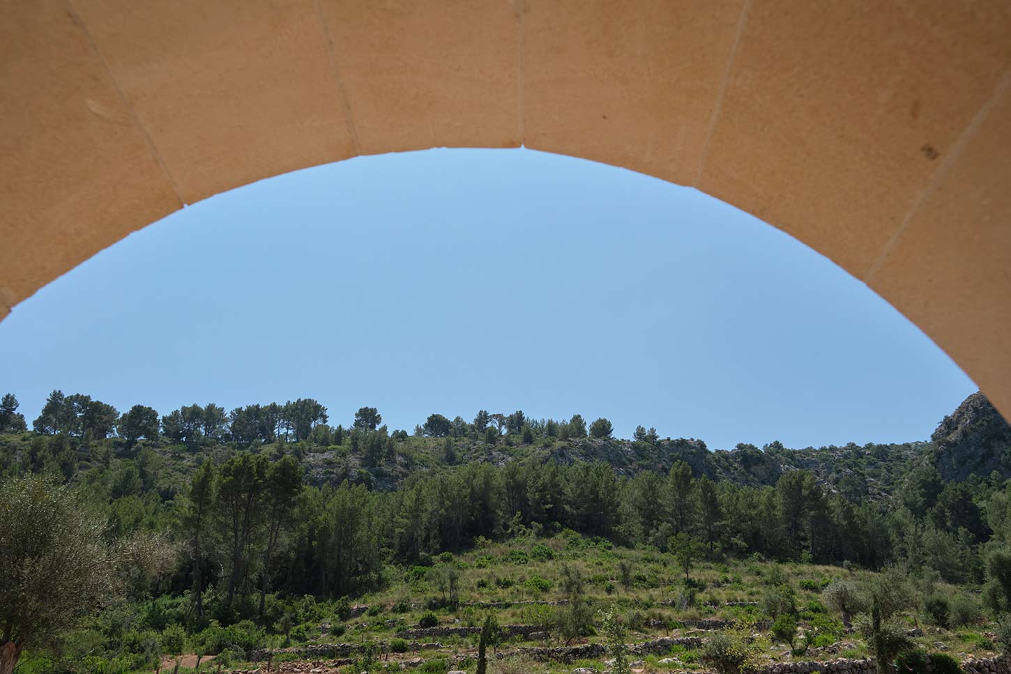 Sandsteinbogen, der eine Berglandschaft mit Pinien und Terrassenfeldern unter klarem Himmel rahmt