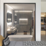 Dormitorio con cabecero rojo y baño en suite visible tras un cristal, con bañera exenta y doble lavabo
