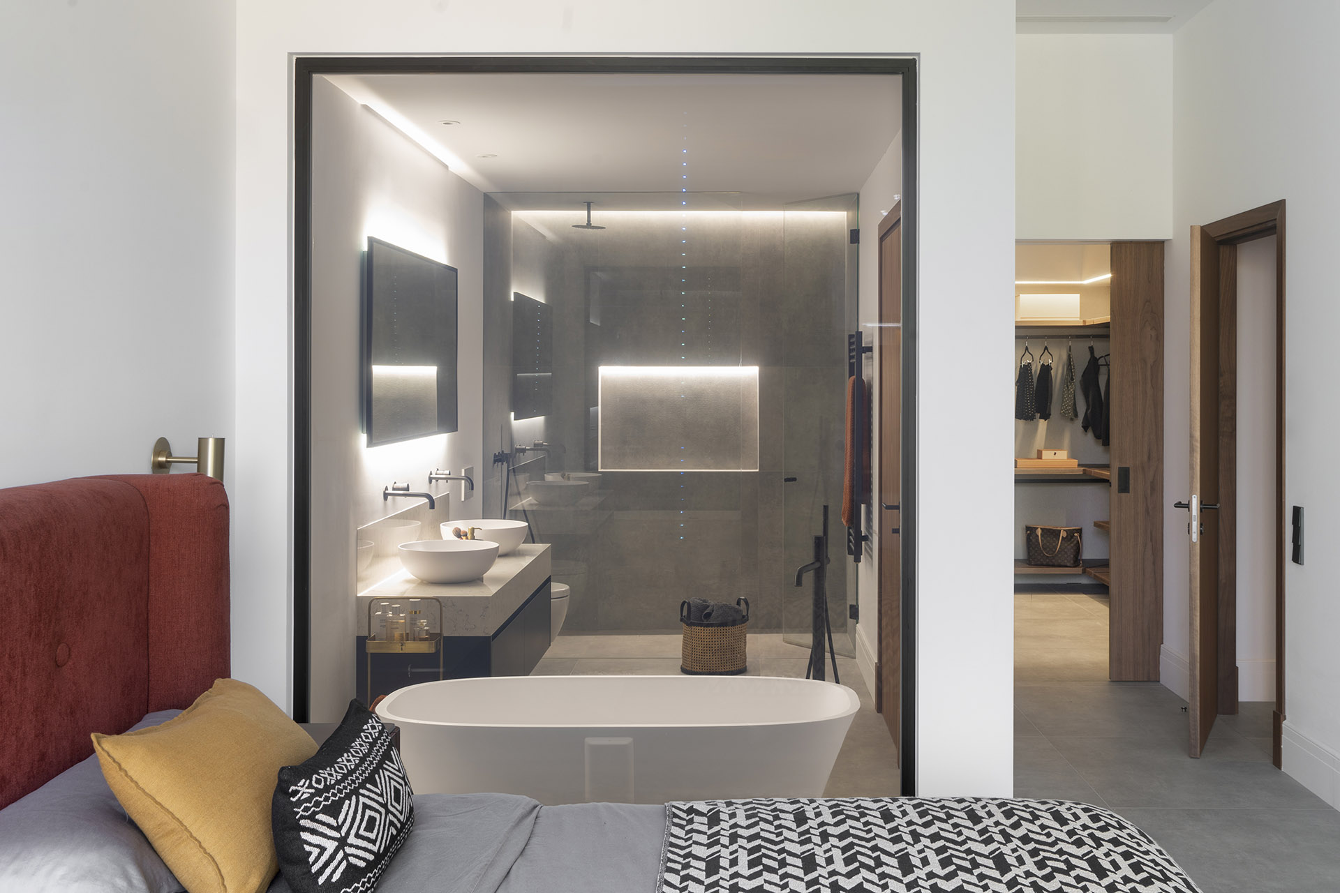 Dormitorio con cabecero rojo y baño en suite visible tras un cristal, con bañera exenta y doble lavabo