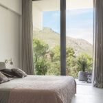 Dormitorio con ventanal de suelo a techo, cortinas de lino y vistas a la montaña entre pinos