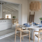 Comedor de terraza cubierta con mesa redonda blanca, sillas de madera y lámpara de fibra abierto a la cocina