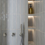 Detalle de ducha con azulejos verticales azul grisáceo, hornacinas iluminadas y grifería de acero