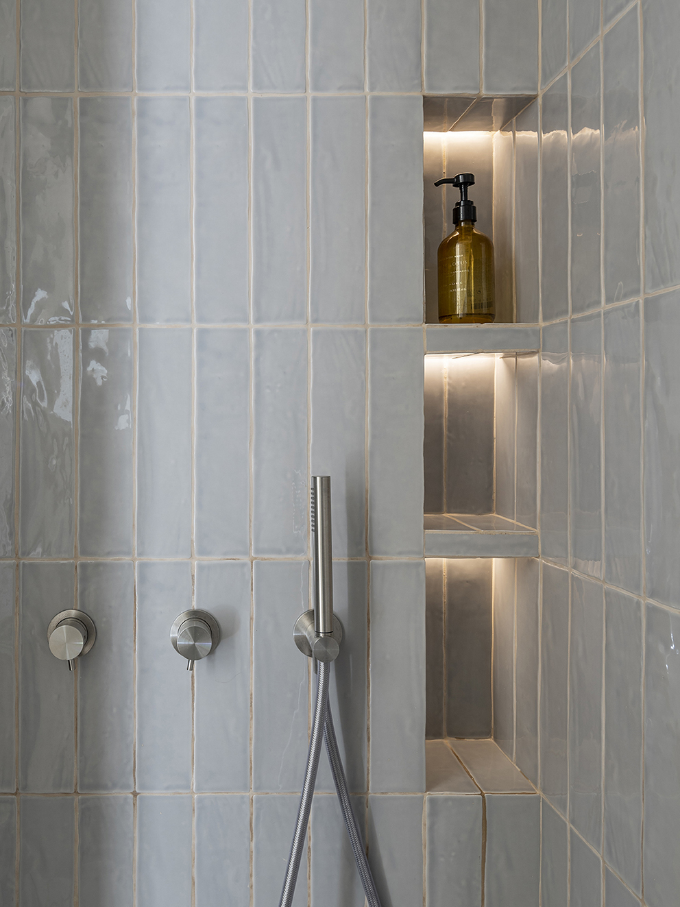 Detalle de ducha con azulejos verticales azul grisáceo, hornacinas iluminadas y grifería de acero
