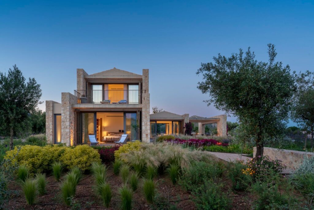 Fachada de la villa Can Pera al anochecer, con piedra, dormitorio iluminado, balcón y jardín con olivos y gramíneas